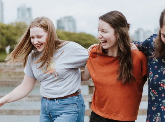 Three women laughing in front of a river, wave goodbye to white marks