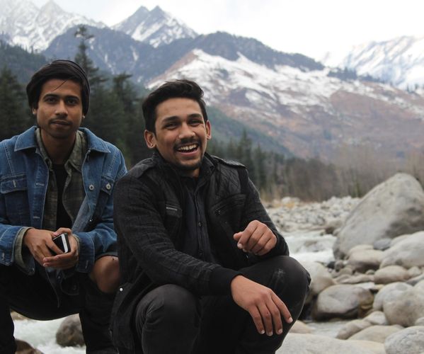 Two men crouching by a river smiling at the bottom of a mountain 