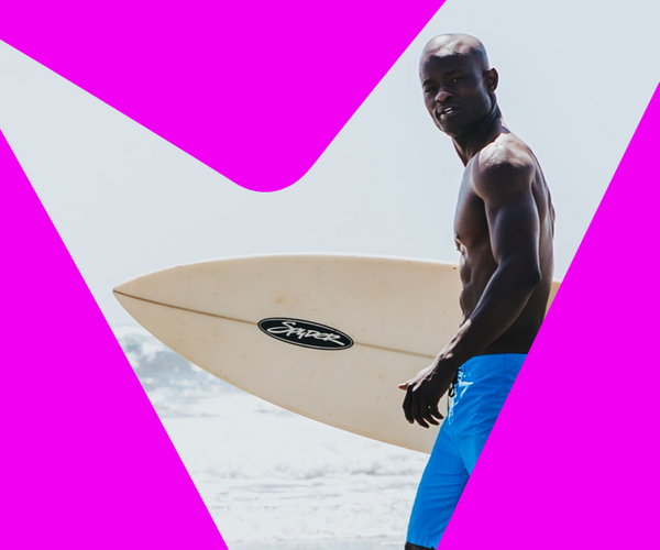 A man in bright blue shorts carries a surfboard along a beach