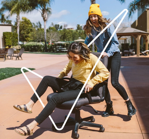 woman in a yellow hat pushing her friend outside on a computer chair. Both girls are laughing and having fun